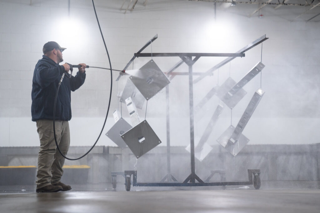 Worker at Biltwood Powder Coating pressure washing metal parts for surface preparation in an industrial facility.
