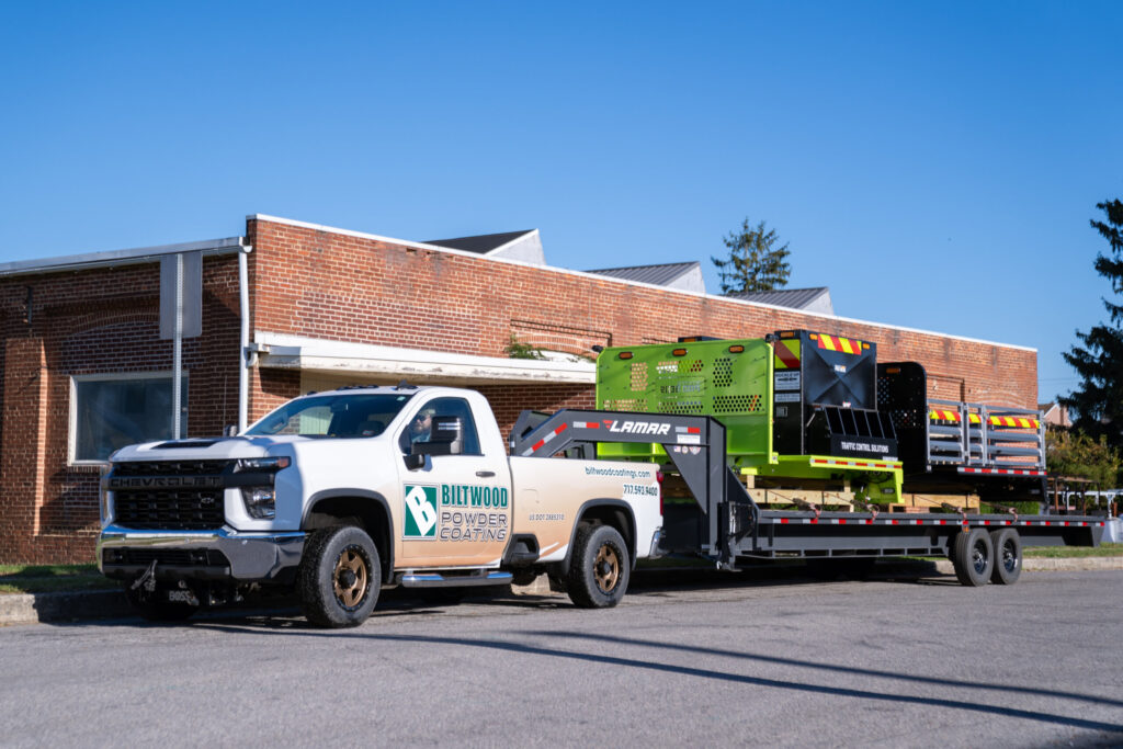 Branded Biltwood Powder Coating truck towing a Lamar trailer with freshly powder-coated industrial equipment outside a brick facility.